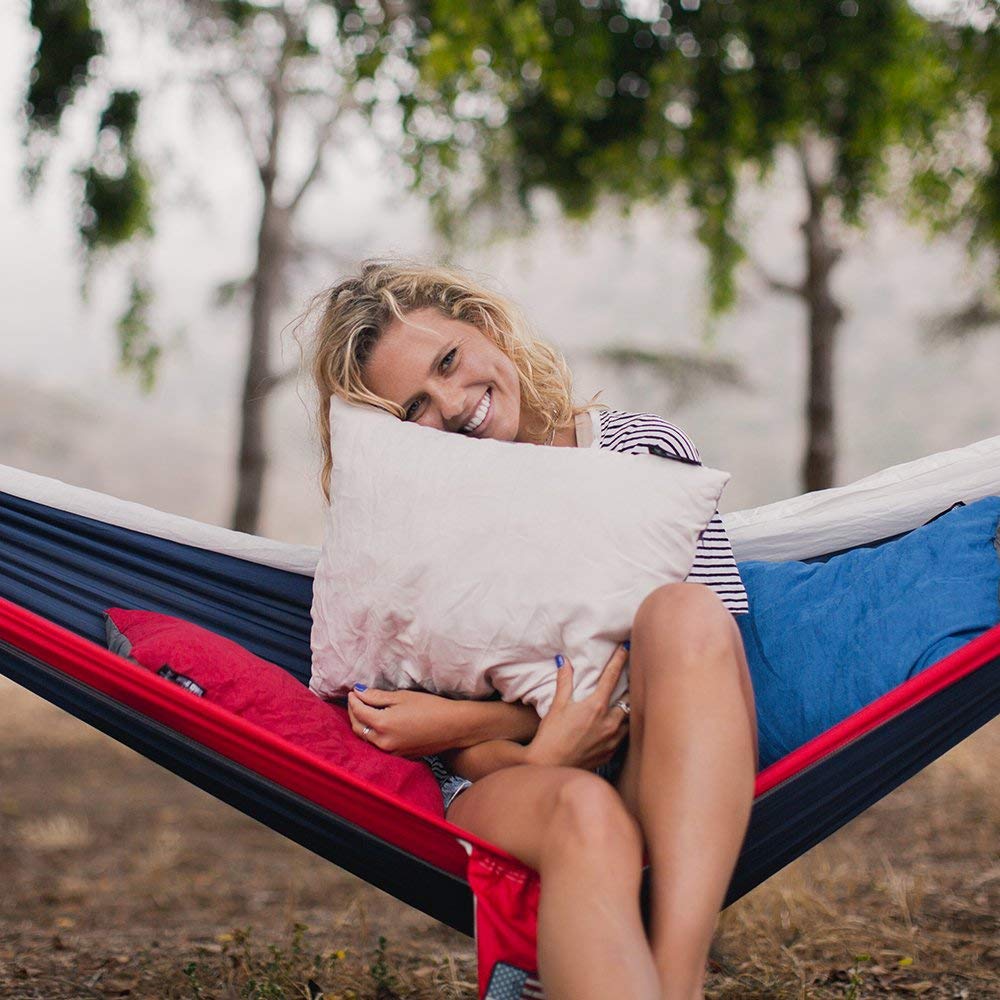 Women holding a camping pillow in a hammock while smiling outdoors.